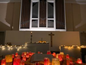 Ein stimmungsvoller Kirchenraum mit einer großen Orgel im Hintergrund und einem zentralen Tisch, auf dem zahlreiche rot und orange leuchtende Kerzengläser eine aufgeschlagene Bibel umrahmen.