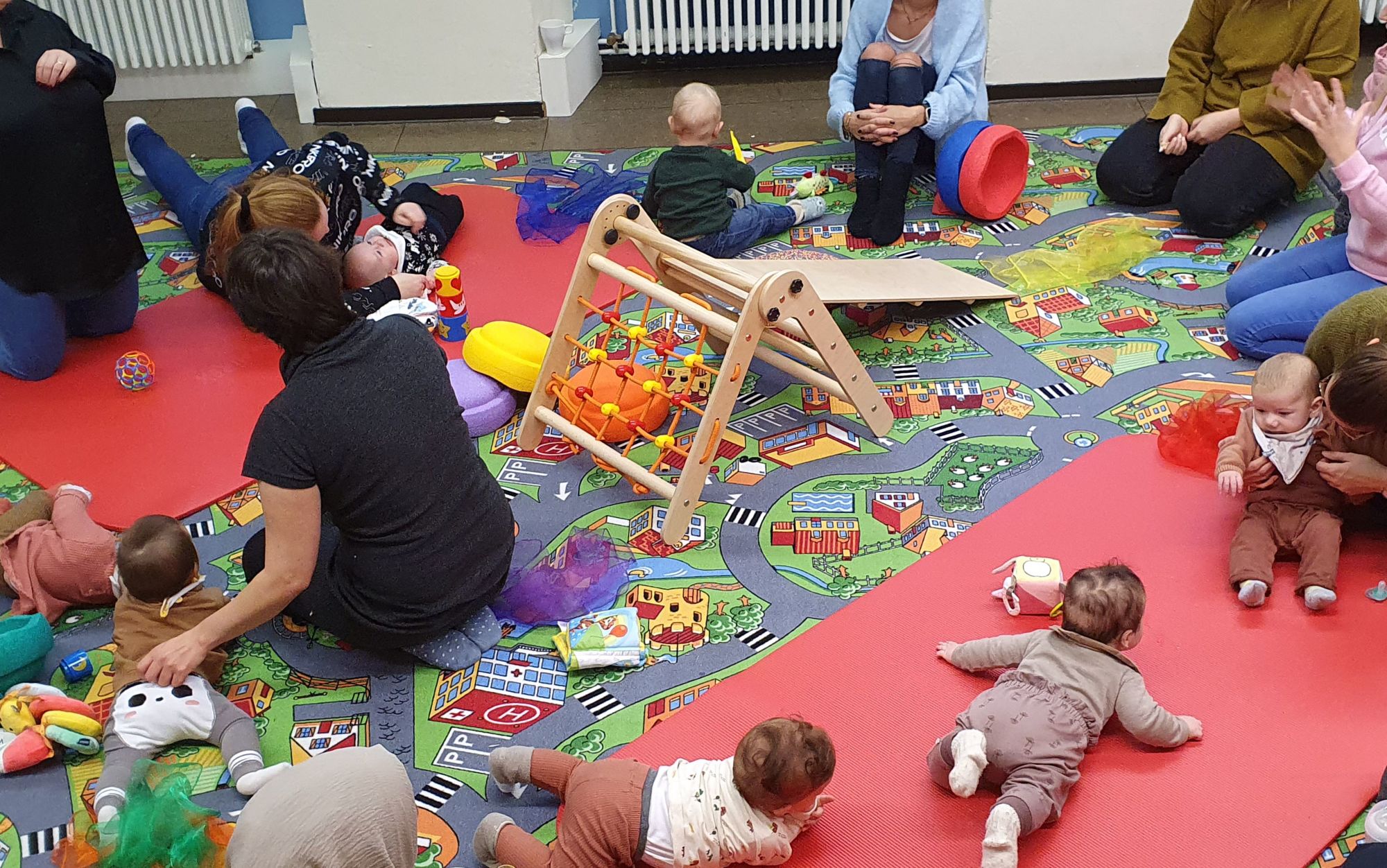 Babys und Eltern in einer Krabbelgruppe mit Klettergerüst und Spielmatten.