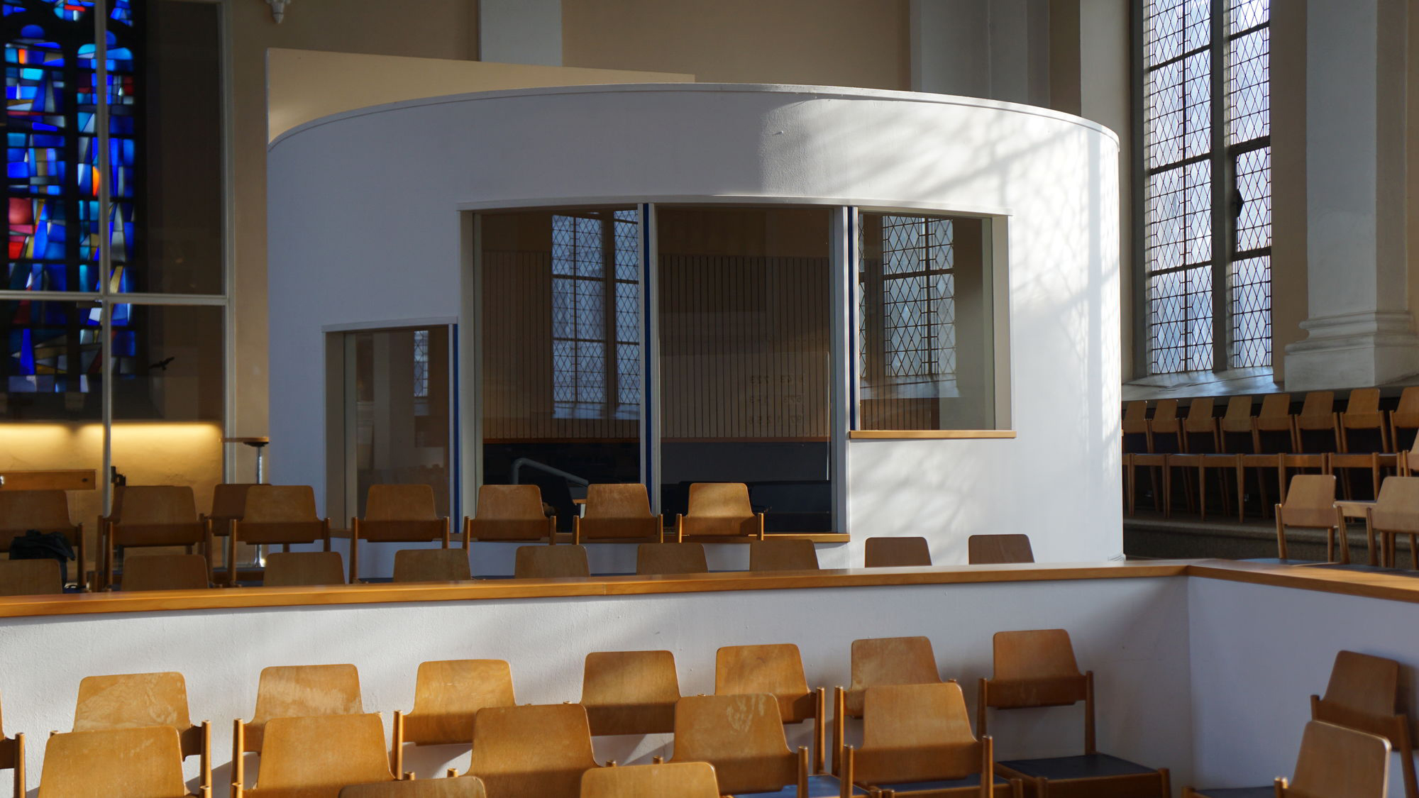 Ein moderner, weißer Rundpavillon aus Glas im Kirchenraum, umgeben von Holzstühlen und traditionellen Buntglasfenstern.