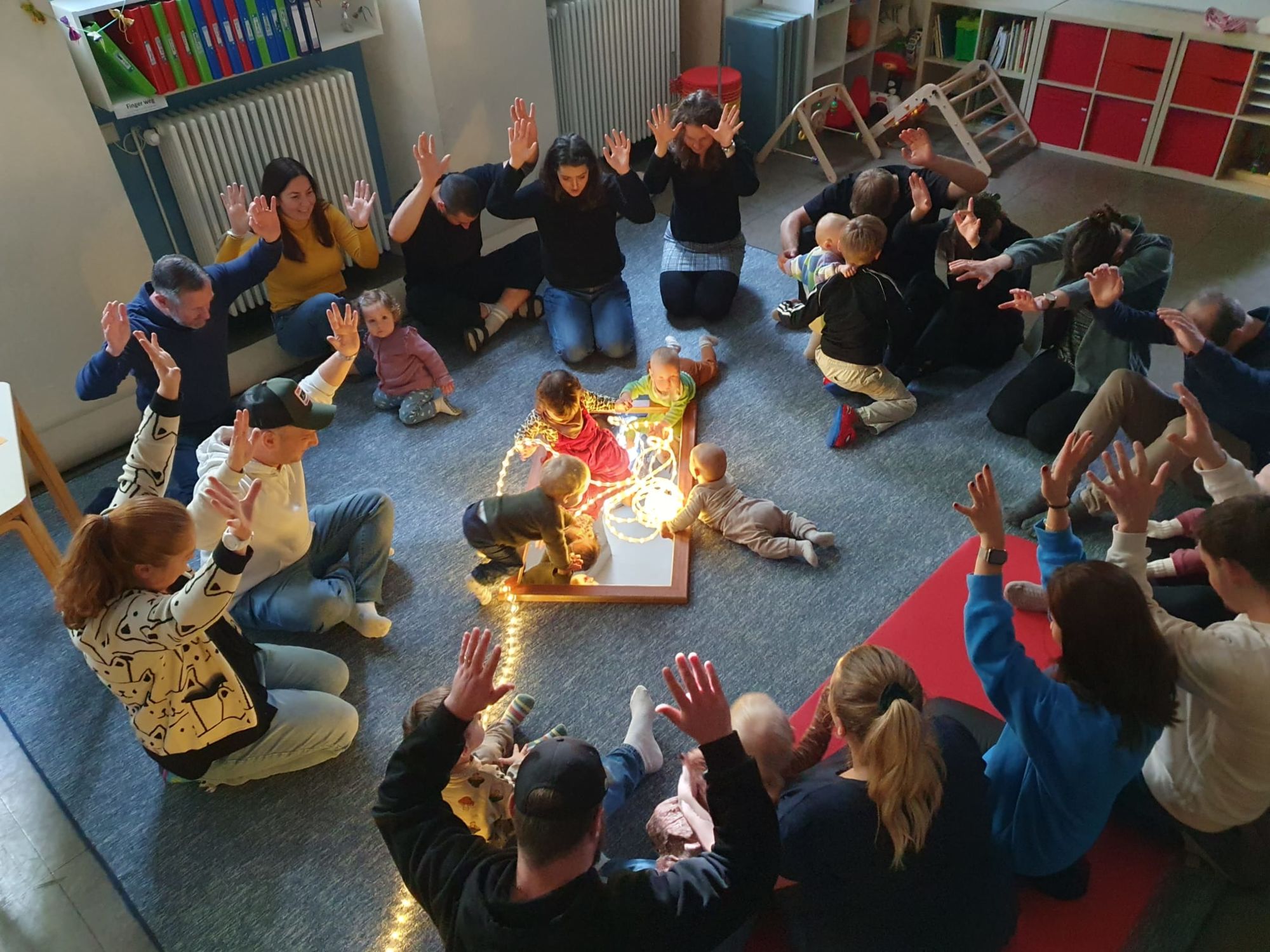 Eltern und Kleinkinder im Kreis um Lichter während einer Krabbelgruppe.